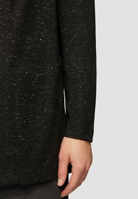 Schwarzes, strukturiertes Langarmshirt mit Sprenkeln, das einen gerundeten Saum und geknöpfte Ärmel hat, teilweise gezeigt mit einer Hand, die den Stoff greift.