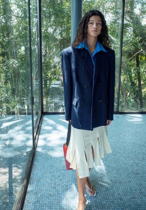 Vrouw met lang krullend haar die een oversized marineblauwe blazer, een blauw overhemd, een witte rok met splitten draagt, een rode tas draagt, staande in een glazen kamer met groen buiten.