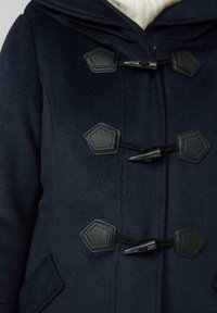 Marineblauer Mantel mit drei schwarzen Knebelverschlüssen und strukturierten Lederflicken, getragen über einem cremefarbenen Strickpullover.