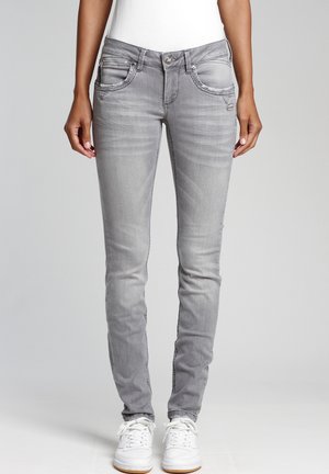 Nainen, jolla on päällään kapeat haalistuneet harmaat farkut, valkoiset tennarit ja valkoinen paita, seisoo yksivärisen harmaan taustan edessä.