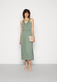 Vestido maxi em tecido verde suave com decote em V, cintura franzida e silhueta fluida. Combinado com sandálias de tiras em tom claro e uma carteira.