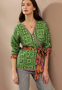 Haut cache-cœur à motif vert avec des bords roses, doté d'une ceinture florale vibrante orange et violette. Manches longues avec poignets froncés.