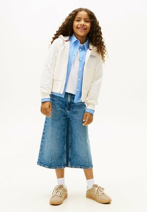 Felpa bianca con zip sopra una camicia a righe blu e bianche, abbinata a una lunga gonna di jeans blu e scarpe da ginnastica beige. Presenta accenti femminili.