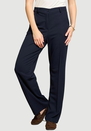 Femme portant un pantalon droit bleu marine, un haut blanc et des chaussures marron à lacets, debout, jambes croisées et mains posées sur les cuisses.