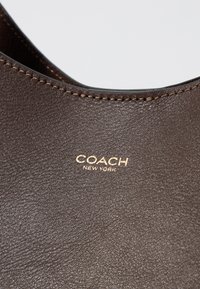 Borsa a spalla in pelle marrone con una superficie liscia, logo dorato in rilievo "COACH NEW YORK" e cuciture marroni a contrasto lungo i bordi.