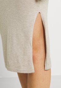 Pletené šaty světle béžové barvy s bočními rozparky, charakterizované hladkou texturou a zakončením lemů. Materiál působí lehce a neformálně.