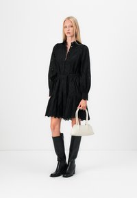 Svart, kne-lengde kjole med lange, puffete ermer, broderte detaljer og innsnevret midje. Kombinert med en kremfarget håndveske og svarte støvletter til kneet.