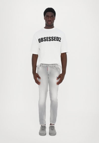 T-shirt blanc avec "OBSESSED2" imprimé en noir, associé à un jean skinny gris clair et des baskets grises, présentant un design classique.