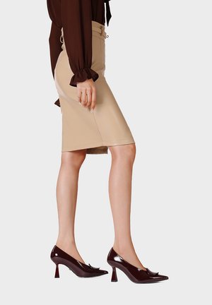 Donna con gonna beige a tubino, camicetta marrone scuro con polsini arricciati e scarpe con tacco alto in vernice marrone scuro, in piedi di lato.