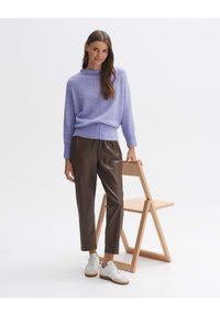 Lichtpaarse gebreide trui, bruine faux leren broek en witte sneakers. Het combineren van texturen met een relaxte pasvorm, staande naast een houten stoel.