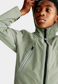 Chaqueta impermeable verde claro con capucha, que cuenta con cremallera frontal y bolsillo lateral con detalle reflectante. Tela suave y con textura.