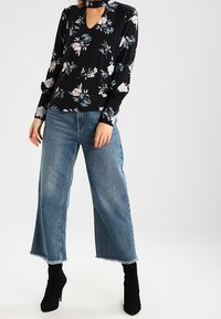 Schwarze Bluse mit Blumenmuster, langen Ärmeln und Schlüsseleckenausschnitt, kombiniert mit blauen Jeans mit weitem Bein und schwarzen Stiefeln mit hohen Absätzen.