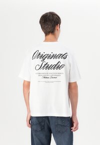 Bílá bavlněná tričko s oversize střihem, s černým textem na zadní straně, včetně nápisu "Originals Studio" a dalších brandingových detailů.