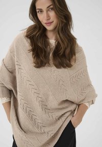 Svetlo bež pletena pončo s chevron vzorcem, širokimi rokavi in teksturiranimi prti. Ima ohlapen, velik fit in mehak material.