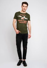 Camiseta verde oliva con un estampado de Wile E. Coyote, jeans negros desgastados y zapatos negros. Diseño casual con mangas cortas y cuello redondo.