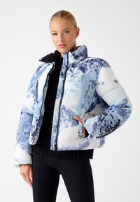 Gevoerde jas in een blauw-wit bergpatroon met hoge kraag, ritssluiting en zijzakken. Een zwarte ribgebreide trui wordt eronder gedragen.