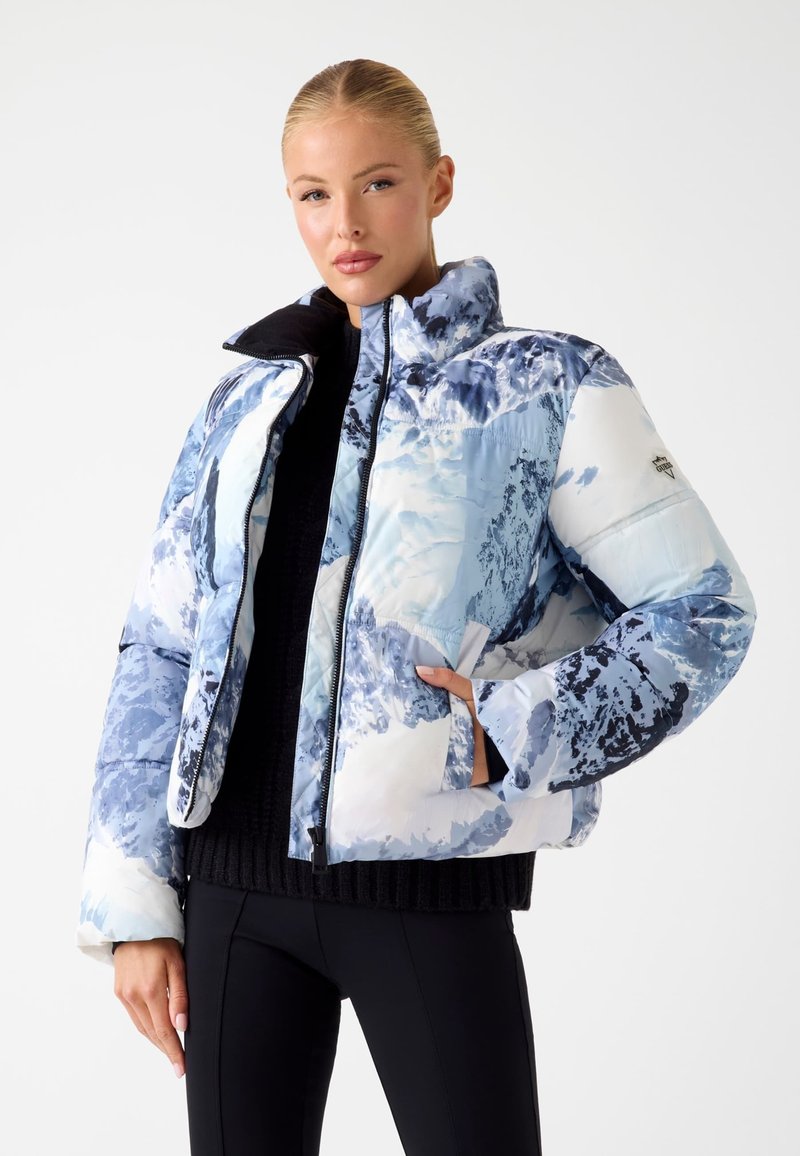 Gevoerde jas in een blauw-wit bergpatroon met hoge kraag, ritssluiting en zijzakken. Een zwarte ribgebreide trui wordt eronder gedragen.