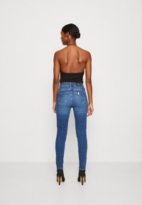 Jeans skinny in denim blu scuro con vita alta, caratterizzati da dettagli minimi e due tasche posteriori; indossati con un top senza spalline e tacchi neri.