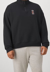 Pullover felpato nero con colletto alto, zip e tre loghi di coccodrillo ricamati in rosso e verde sulla parte anteriore.