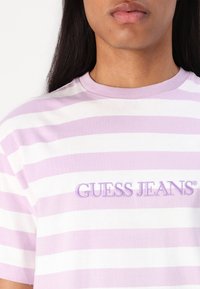 Lilla og hvit stripete bomull T-skjorte med rund hals og brodert "GUESS JEANS"-logo foran. Myk tekstur og avslappet design.