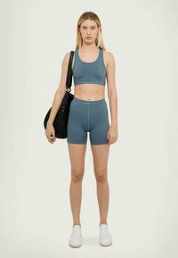 Zilā krāsā pieguļošs divdaļīgs aktīvās apģērba komplekts, kas izgatavots no stiepjama materiāla, satur īsu topu un augstas jostas šortus. Ietver teksturētu melnu somu.