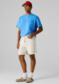 Άνδρας με κόκκινο καπέλο του μπέιζμπολ, μπλε μπλουζάκι, μπεζ σορτς, λευκές κάλτσες και μπεζ παντόφλες στέκεται μπροστά σε γκρίζο φόντο.