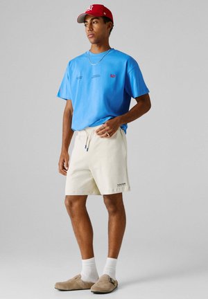 Mężczyzna w czerwonej czapce z daszkiem, niebieskiej koszulce, kremowych szortach, białych skarpetkach i beżowych butach wsuwanych stoi na tle szarego tła.