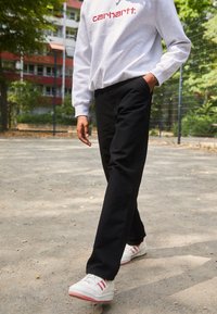 Grauer Hoodie mit rotem Logo, schwarze Hose und weiße Turnschuhe mit roten Streifen. Außenbereich mit Erdboden und grüner Vegetation im Hintergrund.