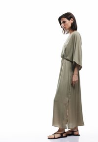 Langes olivgrünes Kleid mit einer lockeren Silhouette, tailliert in der Taille, mit weiten Ärmeln und einem seitlichen Schlitz, kombiniert mit schwarzen flachen Sandalen.