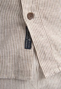 Chemise en tissu rayé beige et blanc avec fermeture par boutons, poche appliquée et étiquette de la marque. Texture douce avec un matériau léger et respirant.