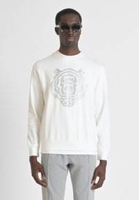 Sudadera blanca con un gráfico de la cara de un tigre en tonos grises, con un cuello redondo y mangas largas. Se llevan pantalones grises ajustados debajo.