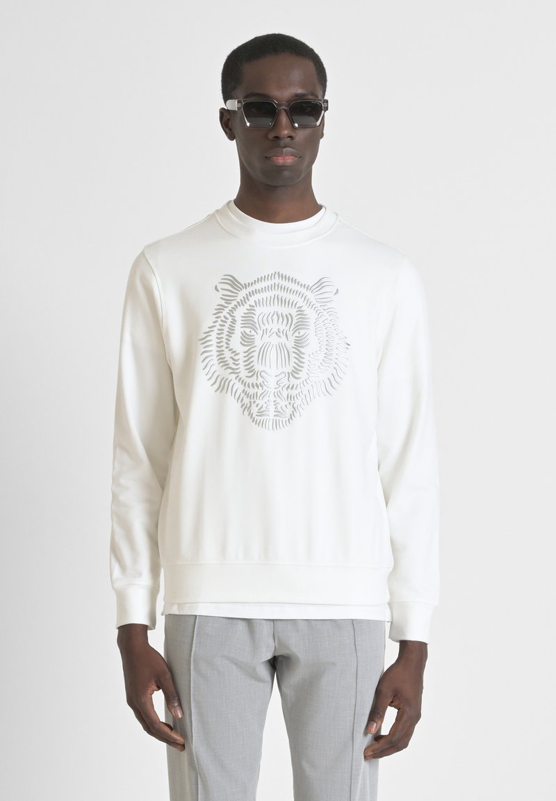 Sudadera blanca con un gráfico de la cara de un tigre en tonos grises, con un cuello redondo y mangas largas. Se llevan pantalones grises ajustados debajo.