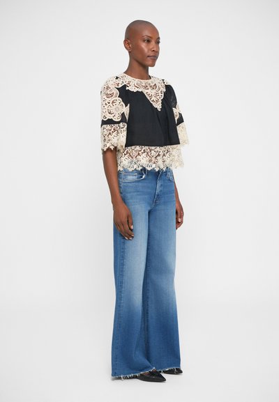 Top in pizzo nero e crema con maniche corte, sovrapposto a un top scuro; abbinato a jeans a gamba larga blu chiaro e scarpe nere a punta.