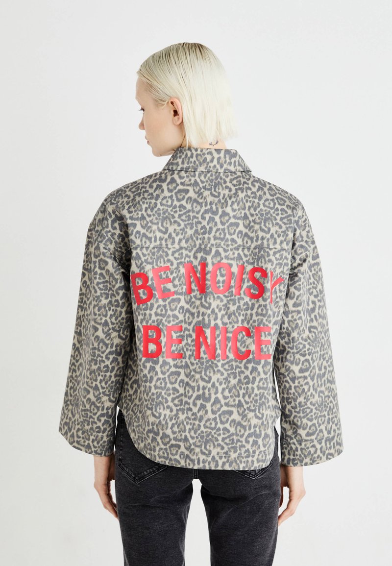 Veste imprimée léopard en tons de gris, avec un texte rouge audacieux "SOIS BRUYANT SOIS AIMABLE" à l'arrière. Fabriquée en tissu doux avec une coupe décontractée.