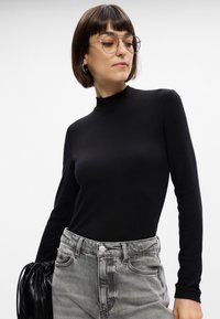 Garšas piedurkņu melna turtlenecka krekls, izgatavots no elastīga auduma, apvienots ar augsto vidukļa pelēko džinsu un melnu maisiņu ar sakrautā dizaina. 