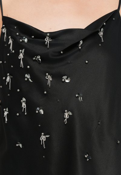 Musta silkki toppi drapeerattulla pääntiellä, jossa on hopeisia helmiä, paljetteja ja koristeita, jotka muistuttavat pieniä hahmoja kankaan pinnalla.