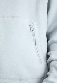 Ljusblå fleecetröja med en dragkedjeförsedd framficka, mjuk textur och synliga sömmar längs kanterna.