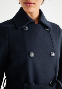 Navy wool-blend coat with a double-breasted front, large buttons, and a belted waist. Features a spread collar and smooth texture.