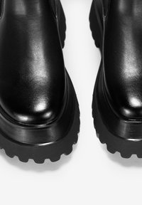 Botines de cuero negro con un acabado brillante, que presentan una suela de goma texturizada gruesa y un diseño de punta redonda.