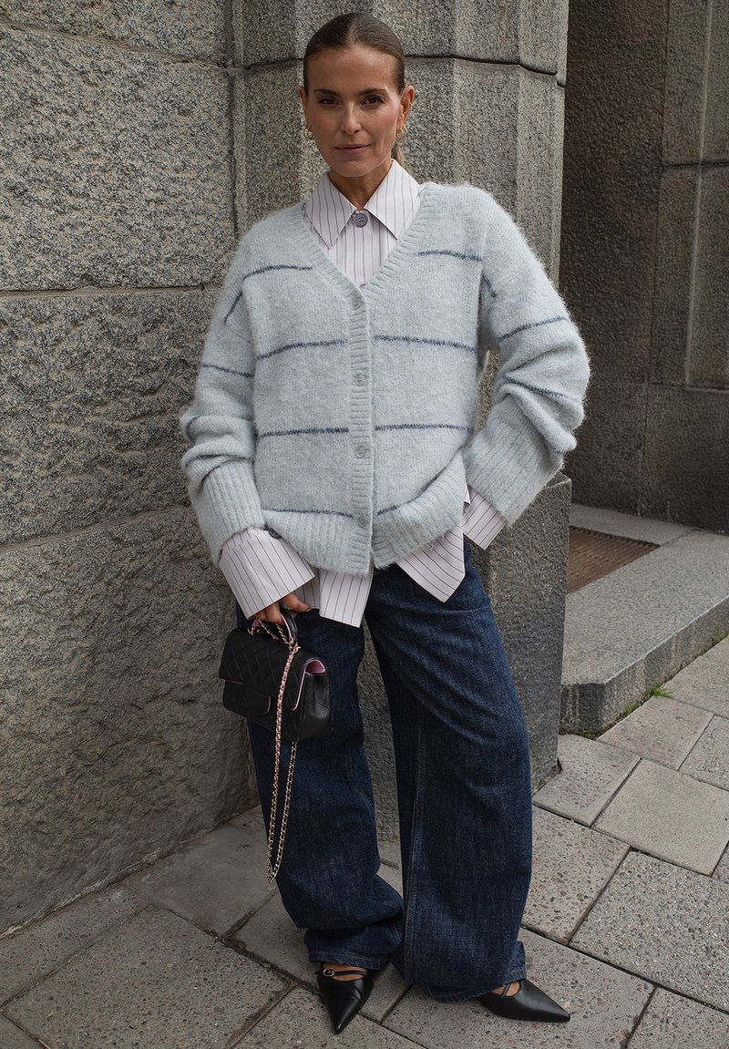 Helles, blaues, flauschiges Strickjacke mit dunklen Streifen, über einem weiß mit vertikalen Streifen versehenen Hemd getragen. Kombiniert mit dunklen weiten Jeans und spitzen schwarzen Schuhen.