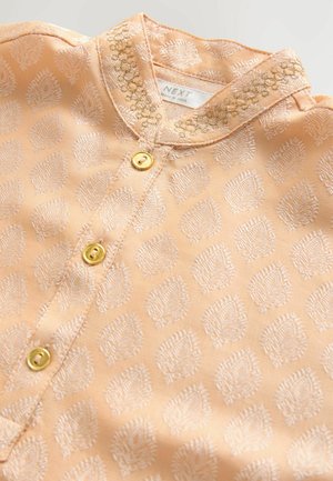 Vaalean persikka värinen kangaspaita, jossa on brodeerattuja lehtikuvioita, kiiltäviä kultanappeja ja koristeellinen kauluksen reuna, jossa on monimutkaista ompelua.