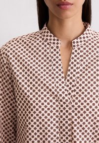 Blouse à manches longues en tissu rose clair, ornée d'un motif géométrique de petites fleurs brunes et d'un col ouvert avec un détail de patte.