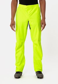 Pantaloni impermeabili giallo neon con una fascia elastica in vita, zip laterale e una texture leggera e lucida. Abbinati a calzature grigie.
