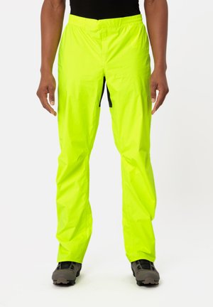 Neon-gelbe wasserdichte Hose mit elastischem Bund, seitlichem Reißverschluss und leichter, glänzender Textur. Kombiniert mit grauem Schuhwerk.