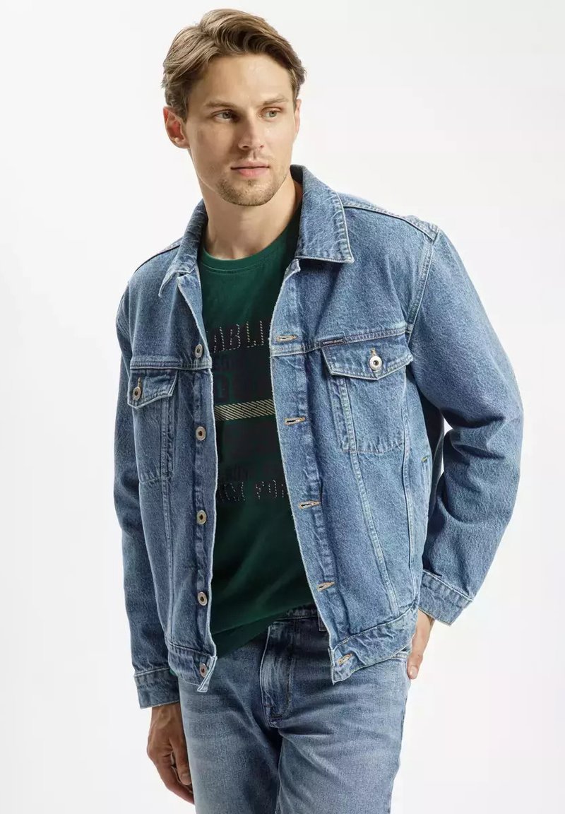Jasnoniebieska kurtka dżinsowa o klasycznym kroju, z zapięciem na guziki, dwiema kieszeniami na piersi i delikatną teksturą. Nosi się ją na ciemnozielonej koszuli.