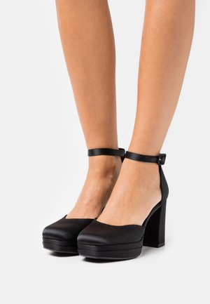 Talons plats noirs avec un bout arrondi, une lanière de cheville et un talon carré. Fabriqués en matériau lisse avec une finition mate.