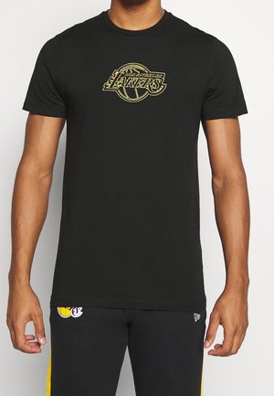 Mężczyzna ubrany w czarny t-shirt z złotym logo Los Angeles Lakers oraz czarne spodenki z emblematem Lakers i żółtymi bocznymi paskami.