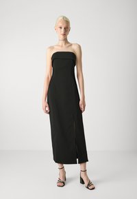 Melna bezsleeve maksidress ar taisnu dekoltē, sānu šķelšanos un gludu tekstūru. Apvienots ar melniem augstpapēžu sandaliem.