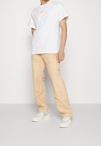 White t-shirt with orange Nike logo, beige cargo pants with pockets, and gray and white sneakers. Simple, athletic design.