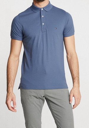 Mann trägt ein eng anliegendes blaues Polohemd mit kurzen Ärmeln und drei Knöpfen sowie graue Hosen, steht vor einem schlichten Hintergrund.